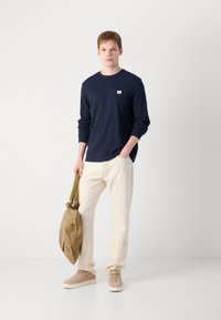 Marineblauwe longsleeve shirt met een klein logo, crèmekleurige jeans, beige sneakers en een tan tas met een groene strap.