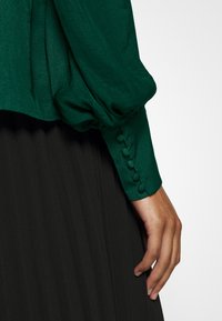 Blusa verde con maniche a sbuffo, realizzata in un tessuto strutturato e con dettagli di polsini con bottoni. Indossata sopra una gonna nera plissè.