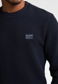 Marineblå sweatshirt lavet af struktureret stof med et honningkage-mønster; har et mærketikette på brystet og ribbet halsudskæring.