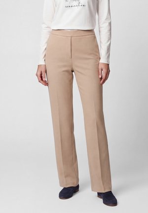 Person wearing beige straight-leg trousers, white long-sleeve top with text, and dark blue loafers, standing against a plain background.