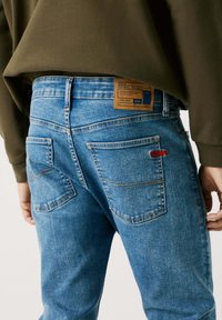 Lyseblå denimjeans med en brun etikett med logo, røde sømdetaljer, og to baklommer med en unik form og design.