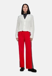White ribbed cardigan with buttons, layered over a black polka dot turtleneck. Red pants with side stripes, paired with chunky black shoes.