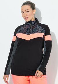 Donna che indossa un giubbotto zip-up nero e grigio con dettagli a zigzag rosa e stampa animale vicino al collo, abbinato a pantaloni rosa acceso.