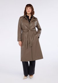 Trench coat lung, cu curea, dintr-un material lucios de culoare verde măslinie, cu guler în formă de V și buzunare laterale, purtat cu pantaloni negri și cizme bej.