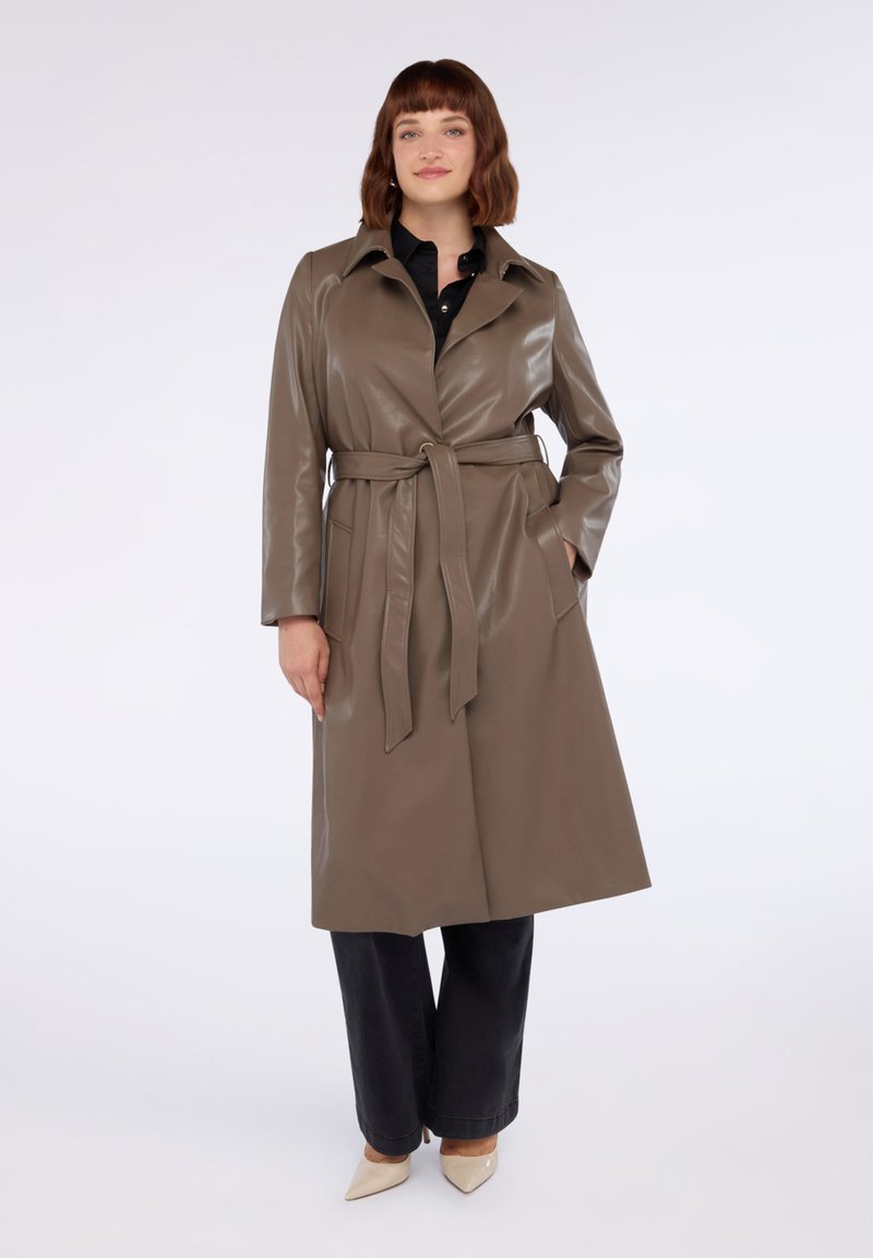 Trench coat lung, cu curea, dintr-un material lucios de culoare verde măslinie, cu guler în formă de V și buzunare laterale, purtat cu pantaloni negri și cizme bej.