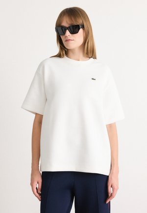 White oversized sweatshirt with short sleeves, featuring a small green logo. Worn with black sunglasses and navy blue pants. Smooth texture.