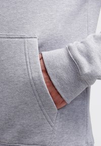Ręka częściowo włożona w przednią kieszeń jasnoszarego sweatshirtu z żebrowanym mankietem i widocznymi szwami.