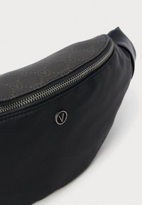 Črna tkaninska torbica za pas s srebrnim zadrgo in temnim vzorčastim poklopcem, z majhnim okroglim kovinskim logotipom s črko "V".