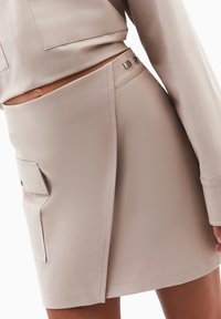 Beige mini nederdel lavet af glat stof, med en sidelomme og metal hardware detaljer. Skræddersyet design med en asymmetrisk søm.