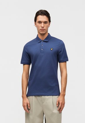 Νέος άνδρας με σκοτεινά μαλλιά, που φοράει μπλε κοντομάνικο πόλο με κίτρινο λογότυπο πουλιού και μπεζ παντελόνια, σε απλό φόντο.