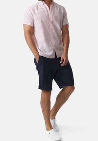 Chemise rose clair à manches courtes et à boutons avec des rayures blanches, associée à un short navy retroussé et des chaussures blanches à enfiler. Tenue d'été décontractée.
