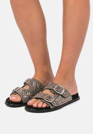 Pies con sandalias negras sin cierre, con dos tiras con hebillas adornadas con tachuelas plateadas y doradas sobre un fondo neutro.