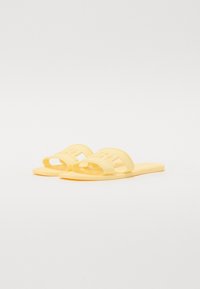 Gula gummislidesandaler med breda remmar som har utklippta bokstäver och en platt sula. Slät yta och minimalistisk design.