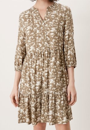 Beige knælængde kjole med hvidt blomsterprint, trekvartærmer, lagde kjoler og en åben V-udskæring båret af en stående person.