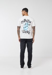 Mand med tatoveringer iført en hvid T-shirt med blå terningegrafik og teksten "feeling lucky", sorte jeans og hvide sneakers, stående med ryggen til.