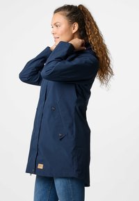 Giacca impermeabile blu navy con cappuccio, tasche laterali e chiusure a scatto. Presenta una vestibilità dritta e una finitura in tessuto testurizzato.