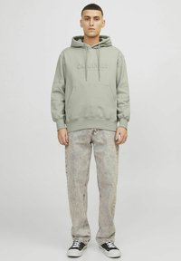 Sudadera con capucha de color verde claro, con bolsillo frontal y logo en relieve "ORIGINALS". Lleva puesto un pantalón vaquero claro con manchas y zapatillas negras.