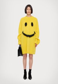 Abito maglione oversize giallo realizzato in materiale morbido, con un design di faccina sorridente nera. Abbinato a una borsa nera strutturata e calze alla caviglia.