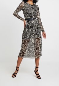 Genomskinlig klänning med långa ärmar i svart och beige med djurmönster, åtdragen i midjan med ett svart bälte, kombinerad med svarta klacksandaler.