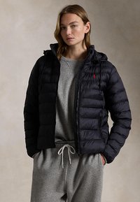 Jonge persoon die een zwarte doorgestikte jas draagt over een grijze trui en bijpassende grijze joggingbroek, handen in de zakken, neutrale achtergrond.