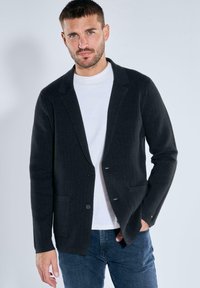 Mann trägt ein dunkles strukturiertes Sakko über einem weißen T-Shirt und blauen Jeans und steht vor einem einfarbigen hellgrauen Hintergrund.