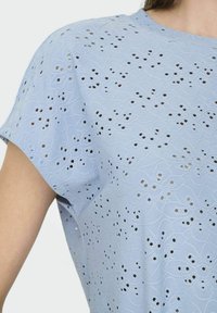 Lyseblå kortærmet top med et laserskåret mønster med blomsterdesign og små huller, lavet af struktureret stof.
