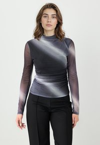 Kobieta z brązowymi włosami do ramion, ubrana w szary, siateczkowy top z długim rękawem o gradientowym cieniowaniu oraz czarne, dopasowane spodnie, stojąca na białym tle.