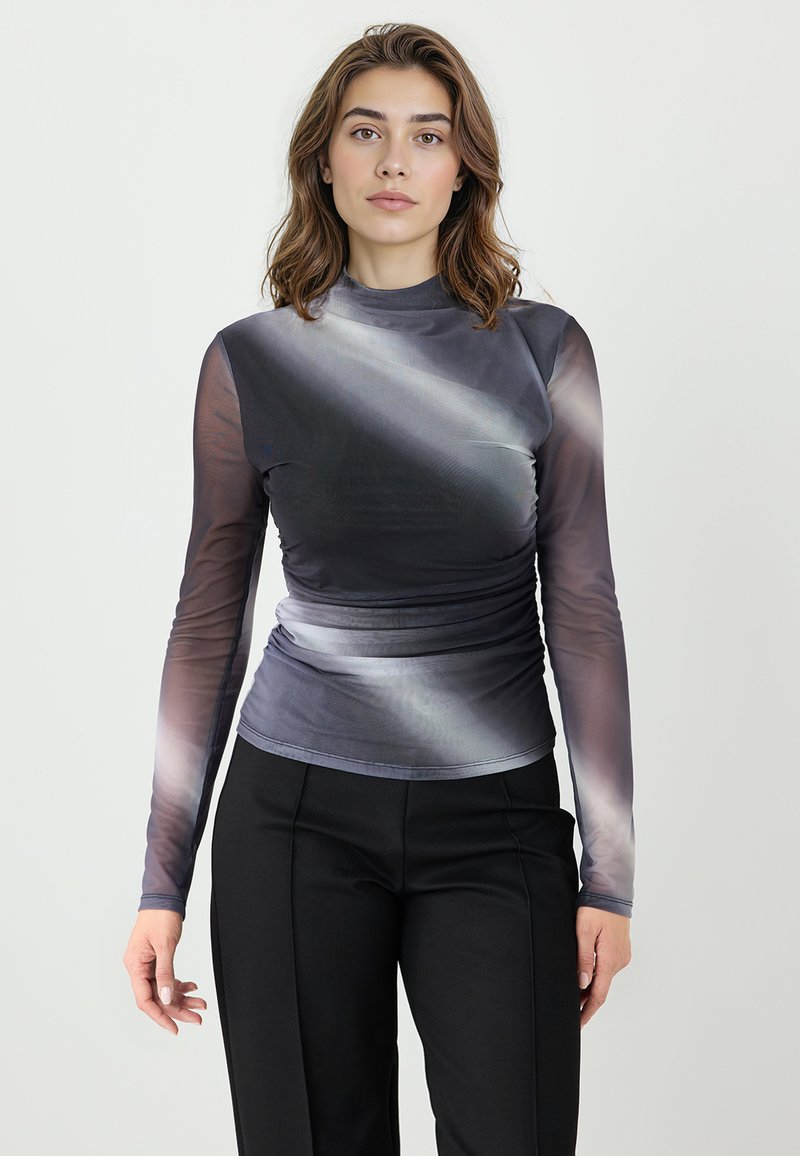 Kobieta z brązowymi włosami do ramion, ubrana w szary, siateczkowy top z długim rękawem o gradientowym cieniowaniu oraz czarne, dopasowane spodnie, stojąca na białym tle.