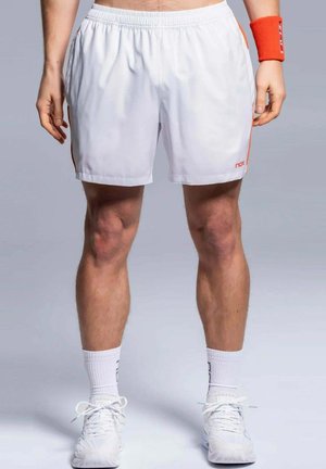 Athlet trägt weiße Sportshorts, weiße Sportschuhe, weiße Socken und ein orangefarbenes Armband, steht vor einem schlichten Hintergrund.
