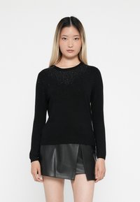 Pull noir à manches longues avec un col rond, orné de sequins sur la poitrine. Porté avec une jupe en simili cuir noire. Texture lisse.