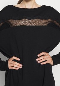 Pull noir à coupe ample, avec une section en dentelle transparente au niveau de la poitrine. Manches longues avec poignets côtelés et texture douce.