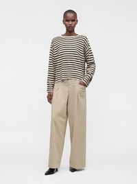 Femme portant une chemise à manches longues beige et noire à rayures, un pantalon large beige et des chaussures pointues noires, debout, une main dans la poche.