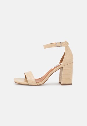 Sandalia de tacón alto en color beige con relieve de cocodrilo, con correa en el tobillo, punta abierta y tacón cuadrado. Textura suave con un acabado brillante.