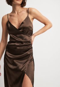 Brązowa satynowa sukienka z dekoltem w kształcie litery V, cienkimi ramiączkami i rozcięciem po boku. Materiał ma gładką fakturę i charakteryzuje się drapowaniem w okolicy talii.