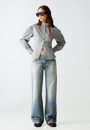 Frau mit großen schwarzen Sonnenbrillen, einer grauen karierten Bluse mit hohem Kragen und Puffärmeln sowie ausgewaschenen, weit geschnittenen Jeans steht mit Händen in der Hüfte.