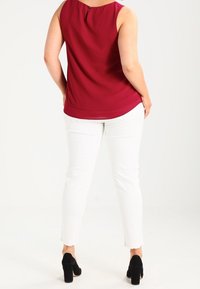 Blouse sans manches bordeaux avec un col rond et un détail dos avec ouverture en forme de goutte, associée à un pantalon skinny blanc et des chaussures à talons noires.
