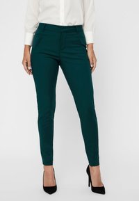 Ciemnozielone, dopasowane spodnie o gładkiej fakturze, z przednimi kieszeniami i średnim stanem. W zestawie z czarnymi wysokimi obcasami.
