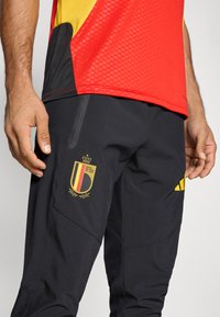 Červený a žltý dres s textúrovaným vzorom, doplnený čiernymi nohavicami s zlatým logom Belgickej futbalovej asociácie a čiernymi zipsami.