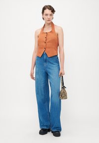 Kvinne med oransje ermeløs skjorte med knapper, vide blå jeans, svarte platåhæler, holder en liten zebra-print håndveske.
