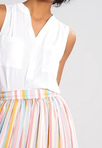 Femme portant un chemisier sans manches blanc avec deux poches sur la poitrine et une jupe plissée à taille haute avec des rayures pastel arc-en-ciel.