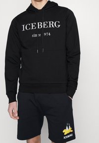 Schwarzer Hoodie mit dem Text "ICEBERG seit 1974", verstellbarem Kordelzug für die Kapuze und Kängurutasche; kombiniert mit dunklen Shorts, die eine Cartoonfigur zeigen.