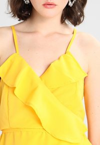 Gelbes ärmelloses Kleid mit rüschigem V-Ausschnitt. Das leichte Gewebe, die glatte Textur und die taillierte Passform betonen die Silhouette.