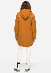 Lång, huvad kappa i bränd orange fleece med textilstruktur och kviltade axelaccentueringar. Bärs över vita byxor och kraftiga stövlar med svart sula.