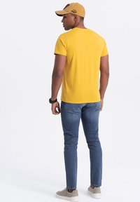 Gul bomulds t-shirt med rund hals, parret med mørkeblå slim-fit jeans. En tan kasket med et logo bæres bagfra. Grå sneakers fuldender looket.