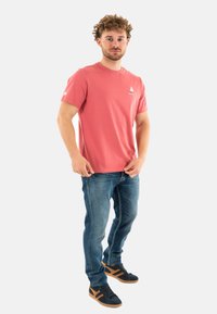 Camiseta de algodón rosa claro con mangas cortas, que presenta un pequeño logo de un velero. Combinada con unos vaqueros de mezclilla azul y zapatillas oscuras.