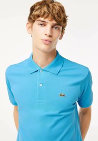 Vaaleansininen polo-paita, joka on valmistettu teksturoidusta kankaasta. Puserossa on klassinen kaulus sekä pieni vihreä krokotiililogo vasemmassa rinnassa.