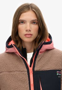Brown fuzzy fleece jacket with a navy panel. Features a pink hood, bright zipper, and logo patch. Comfortable and warm design.