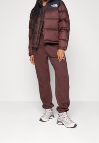 Doudoune bordeaux à col noir, associée à un pantalon de survêtement assorti. Des chaussures de sport grises et violettes complètent la tenue.