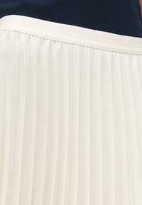 Fustă plisată confecționată dintr-un material alb, texturat, cu linii verticale, asortată cu un brâu solid de culoare albastră închisă și detalii orizontale subtile.