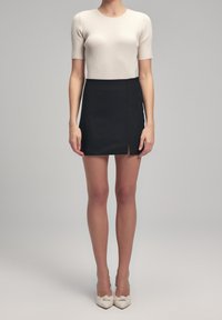 Přiléhavý ribkovaný béžový top, černá mini sukně s předním rozparkem a bílé boty s detailním uzlem. Hladké textury a kontrastní barvy.